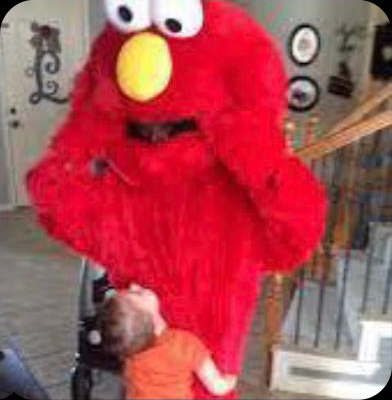A person in an Elmo costume being hugged by a small child, inside what is clearly someone’s house
