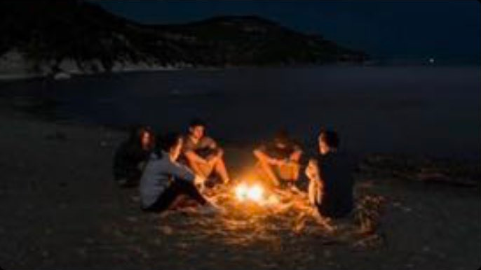 Five people sitting around a campfire on a beach in the dark.
