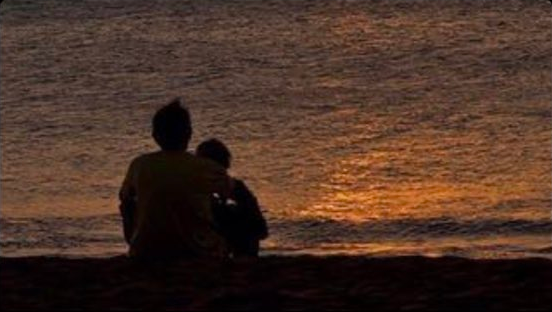 Two people sitting together by a body of water, silhouetted against the sunset’s light. One is leaning on the other.